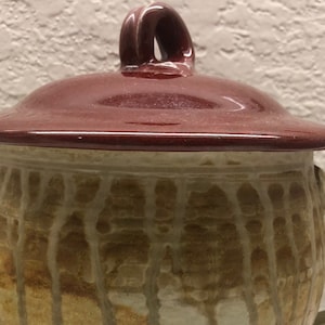 Op de afbeelding: Een keramische mok met een deksel. De mok is bruin en wit met een druppelglazuur. Het deksel is dieprood.