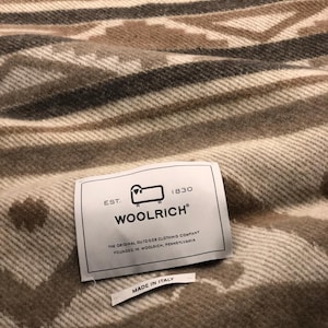 May include: A close-up of a woven blanket with a geometric pattern in shades of beige, brown, and gray. The blanket has a tag that reads "WOOLRICH" and "MADE IN ITALY".