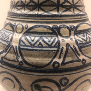 May include: A ceramic vase with a beige base color featuring hand-painted dark blue geometric patterns. The design includes a grid pattern at the neck, circular motifs with internal zig-zag lines, triangles, and swirling scrollwork near the base.