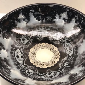 May include: A round glass bowl featuring a black and white lace-like pattern on the interior surface. A small, circular, cream-colored paper doily with a delicate die-cut edge is centered at the bottom of the bowl, contrasting with the dark design.