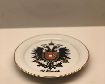 May include: A white decorative plate with a gold rim. The plate features a detailed emblem with a double-headed eagle, a shield, and the text "Alt Osterreich" in black.