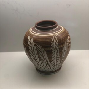 Puede incluir: Un jarrón de cerámica marrón con diseños de hojas estilizadas blancas. El jarrón tiene un cuerpo redondeado y un labio ligeramente acampanado.