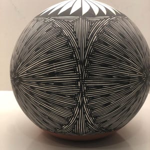 Puede incluir: Una vasija de cerámica esférica con un patrón dibujado a mano en blanco y negro de líneas radiantes y formas geométricas. La sección superior muestra un diseño de pétalos blancos sobre fondo negro, mientras que la base es de color terracota.