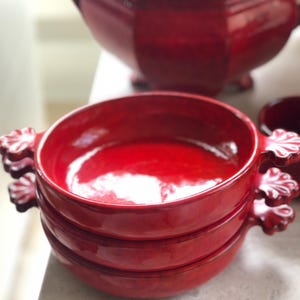May include: A stack of three round, bright red ceramic bowls with glossy finishes. Each bowl features two small, decorative handles on opposite sides, shaped like stylized shells or fans. The bowls have a consistent, vibrant red color.