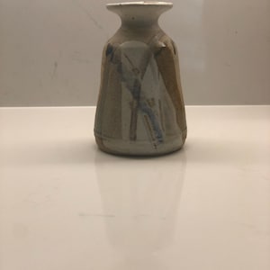 May include: A small, hand-thrown ceramic vase with a wide mouth and a narrow neck. The vase is decorated with brown, white, and blue glaze, creating a unique and abstract pattern.