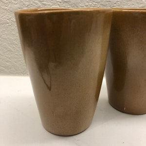 Pode incluir: Duas canecas de cerâmica marrom com acabamento liso e brilhante. As canecas são altas e de forma cilíndrica.