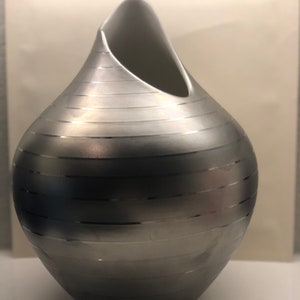 May include: A silver ceramic vase with a unique, asymmetrical design. The vase has a smooth, glossy finish and features horizontal lines.