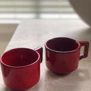 May include: Two matching ceramic mugs with a vibrant red glaze and a darker, mottled finish near the rims. Each mug features a cylindrical body and a distinct, angular square-shaped handle extending from the side, providing a sturdy grip.