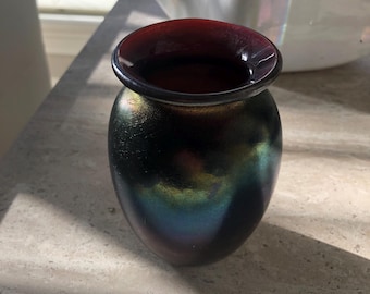May include: A small, dark vase with a rounded body and a flared, reddish-brown rim. The vase has an iridescent finish, displaying hues of green, blue, and gold. The vase is sitting on a light-colored surface.