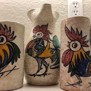 Może przedstawiać: Zestaw trzech ceramicznych wazonów z motywem koguta. Wazy są białe z fakturowaną powierzchnią i przedstawiają kolorowe ilustracje kogutów. Największy wazon to dzbanek z dzióbkiem.