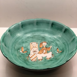 Può includere: Una ciotola in ceramica turchese con bordo smerlato. La ciotola presenta un'illustrazione dipinta a mano di due figure in abiti tradizionali. Le figure sono sedute e la ciotola ha una finitura lucida.
