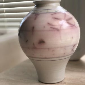 Puede incluir: Un jarrón de cerámica hecho a mano con un cuerpo bulboso y una base acampanada. El jarrón presenta una combinación de colores rosa claro y blanco con acentos morados. Líneas horizontales y patrones abstractos decoran la superficie.