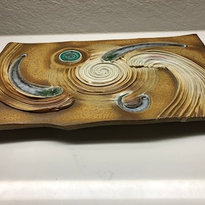 May include: A rectangular ceramic wall hanging with a brown background and white, green, and blue abstract designs. The design features a spiral, curved lines, and circles.