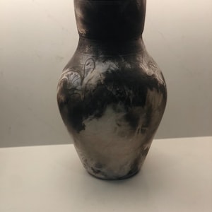 May include: A ceramic vessel with a lid featuring a rounded body and a narrow neck. The surface displays a mottled, smoky pattern in shades of charcoal black, gray, and off-white, with faint, etched linear markings across the exterior finish.
