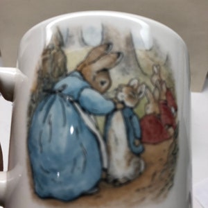 May include: White ceramic mug featuring a scene from a children's story. The illustration depicts a rabbit in a blue dress and a rabbit in a blue jacket, with other rabbits in the background. The mug has a curved handle.