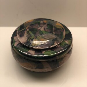 以下が含まれることがあります： 蓋付きの丸い陶器製の小物入れ。箱と蓋には、緑、茶色、黄色の葉の形が模様として描かれています。箱は濃い緑色の縁と薄茶色のベースを持っています。