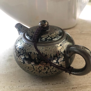 Puede incluir: Una pequeña tetera de cerámica redonda con un patrón de esmalte moteado en plata metálica y negro. La tetera tiene una tapa a juego con un pequeño pomo, un asa curva y un pico corto. Un cordón marrón conecta el pomo de la tapa con el asa.
