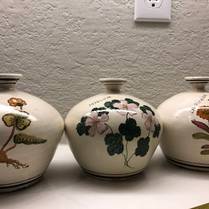 May include: Three white ceramic vases with floral designs. The vases are decorated with hand-painted flowers and leaves. The vases are labelled "Marlyn", "La Ficolosa", and "La Ficolosa".