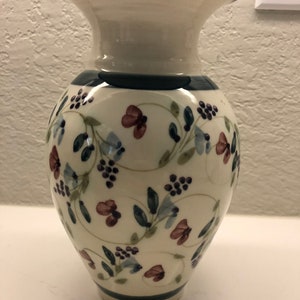 Puede incluir: Un jarrón de cerámica blanco con un patrón floral azul y verde. El jarrón tiene una boca ancha y una base estrecha.