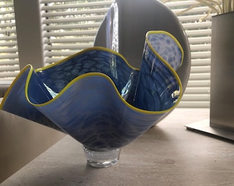 May include: A decorative glass bowl with a flowing, wave-like design. The bowl features shades of blue with yellow trim and sits on a clear, textured base. The bowl's design is reminiscent of ocean waves.