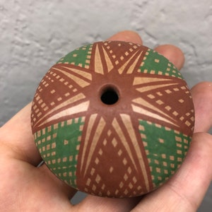 Puede incluir: Un pequeño cuenco redondo de cerámica marrón con un diseño geométrico verde y marrón. El cuenco tiene un pequeño agujero en el centro.