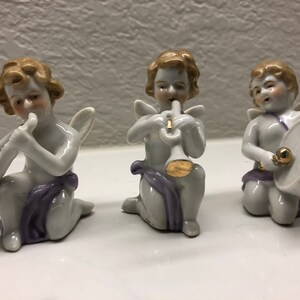 Op de afbeelding: Drie porseleinen cherubijnbeeldjes die muziekinstrumenten bespelen. De cherubijnen zijn wit met gouden accenten en paarse sjerpen. Eén cherubijn speelt saxofoon, een ander trompet en de derde trommel.
