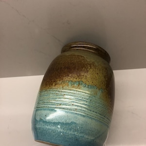 Könnte beinhalten: Eine handgefertigte Keramikvase mit rundem Körper und schmaler Öffnung. Die Vase zeigt einen Farbverlauf, der von Dunkelbraun oben nach Türkisblau unten verläuft, mit einem gelb-grünen Band dazwischen. Die Oberfläche weist horizontale strukturierte Linien auf.