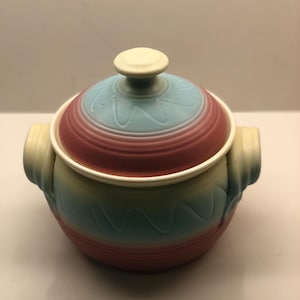 Puede incluir: Una cazuela de cerámica con tapa y asas. La tapa es azul claro y granate, con un diseño ondulado. El cuerpo de la cazuela tiene un degradado de colores, incluyendo azul, amarillo y rojo.