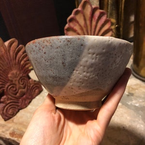 Puede incluir: Un pequeño cuenco de cerámica gris y blanco con un esmalte moteado. El cuenco tiene una forma redondeada y un borde ligeramente acampanado.
