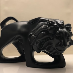 以下が含まれることがあります： しゃがんだポーズのライオンの黒い抽象彫刻。彫刻は滑らかでマットな素材で作られており、スタイリッシュでモダンなデザインです。