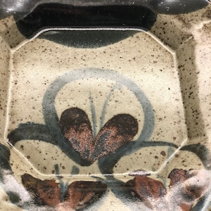 Może przedstawiać: Kwadratowy talerz ceramiczny z jasnobrązową glazurą i ciemnoniebieskim i brązowym wzorem kwiatowym. Talerz ma lekko uniesiony rant i fakturowaną powierzchnię.