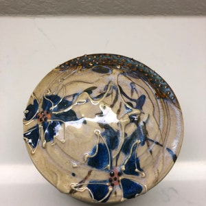 May include: A ceramic lid with a cream base and blue and brown floral design. The lid has a textured rim with gold accents.