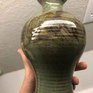 Könnte beinhalten: Eine grüne Keramikvase mit braunen und goldenen Akzenten. Die Vase hat einen breiten Boden und einen schmalen Hals.