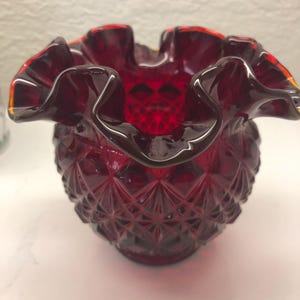 May include: A red glass vase with a ruffled rim and a diamond pattern. The vase is made of clear glass with a red tint.