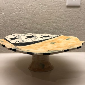 May include: A ceramic cake stand with a white base and a scalloped top. The top is decorated with black and white abstract designs and a yellow and green glaze.