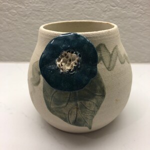 Puede incluir: Un pequeño jarrón de cerámica blanca con un diseño de flor azul y hoja verde. El jarrón tiene una forma redondeada y un acabado liso y mate.