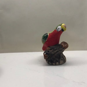 Può includere: Una piccola statuetta di pappagallo in ceramica dipinta di verde, rosso e giallo, posata su un tronco marrone.