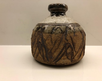 May include: A ceramic vase with a rounded body and a small neck. The vase has a textured, brown surface with dark, linear patterns. The upper section is a lighter colour, and the neck is dark brown. The vase is likely handmade.