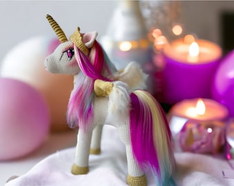 May include: A white crocheted unicorn with a pink, white, and blue mane and tail. The unicorn is wearing a gold crown and has pink eyes. The unicorn is sitting on a white surface with a pink balloon and candles in the background.