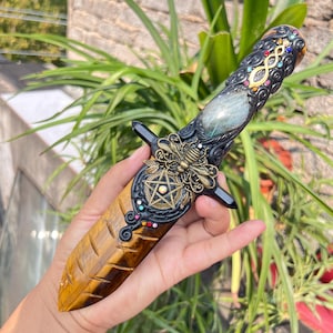 May include: Ornate dagger with a tiger's eye blade and a detailed handle. The handle features a large, oval gemstone, a brass bee, and a pentagram. The hilt is black with colorful accents. The blade is a warm, golden brown color.