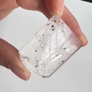 May include: A clear, rectangular crystal with black inclusions. The crystal is held in a hand, showcasing its polished facets and internal patterns. The transparent stone displays a mix of light and dark elements.