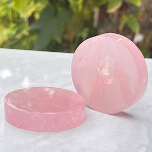 May include: Two polished rose quartz plugs. One is a flat disc, the other is a rounded shape with a groove. Both are a soft pink colour and appear smooth. The background is blurred green foliage.