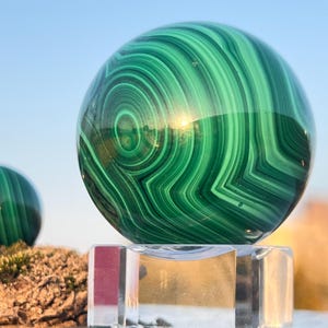 Può includere: Una sfera di malachite lucidata con motivi a spirale di verde scuro e chiaro. La sfera è posta su un supporto rettangolare trasparente, che riflette la luce solare. Lo sfondo è un cielo azzurro, che mette in risalto la bellezza naturale della pietra.