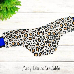 May include: A white fabric dog collar with a leopard print pattern and blue trim. The collar has a black and blue stripe on the left side. "Many Fabrics Available" is written in black text at the bottom of the image.