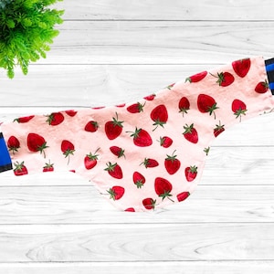 May include: A pink fabric bandana with a strawberry print and blue and black trim. The bandana is folded in half and lying on a white wooden surface.