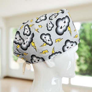 May include: A gray surgical scrub cap featuring a pattern of cartoon storm clouds with angry faces and yellow lightning bolts. The cap has a white tie closure at the back. The cap is designed to be worn in a medical setting.