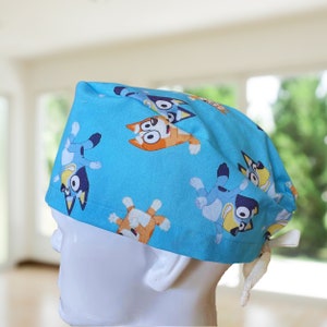 May include: Blue surgical scrub hat with a repeating pattern of cartoon dogs in various poses. The dogs are blue, orange, and brown with white accents. The hat has a white drawstring.