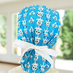 May include: A blue surgical scrub hat with a white pattern of cartoon vertebrae with different facial expressions. The hat has white ties that can be tied at the back of the neck.