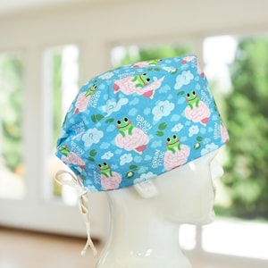 May include: A light blue scrub cap featuring a repeating pattern of cartoon frogs sitting on pink brains, with white clouds and question marks. The words "BRAIN FROG" are printed on the cap. The cap has a white tie closure.
