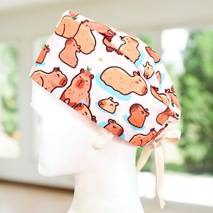 May include: A white surgical cap featuring a repeating pattern of cartoon capybaras in various poses. The capybaras are primarily shades of brown and peach, with some in blue water. The cap has a cream-colored tie.